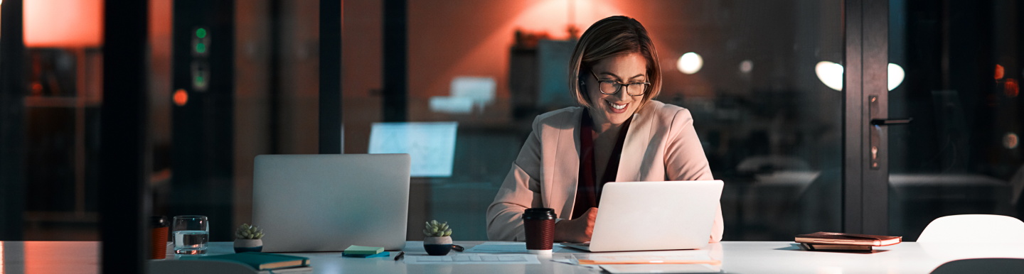Banner central con la imagen de una mujer sonriente vestida con un blazer claro, trabajando de noche en una oficina moderna, mirando una notebook. Hay una taza, un vaso de agua, plantas pequeñas y otra laptop sobre la mesa.