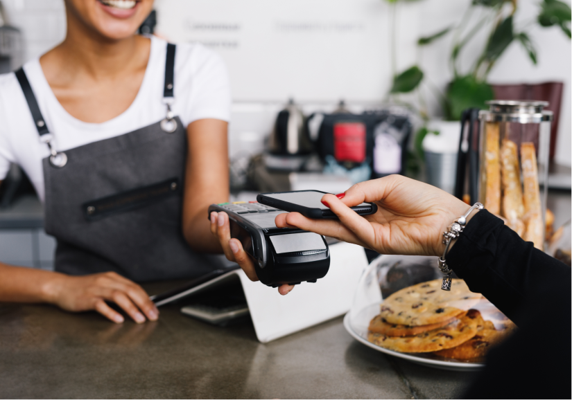 Cliente realizando un pago sin contacto con el celular en una terminal, con una empleada sonriente al fondo en una cafetería con galletitas exhibidas en el mostrador.