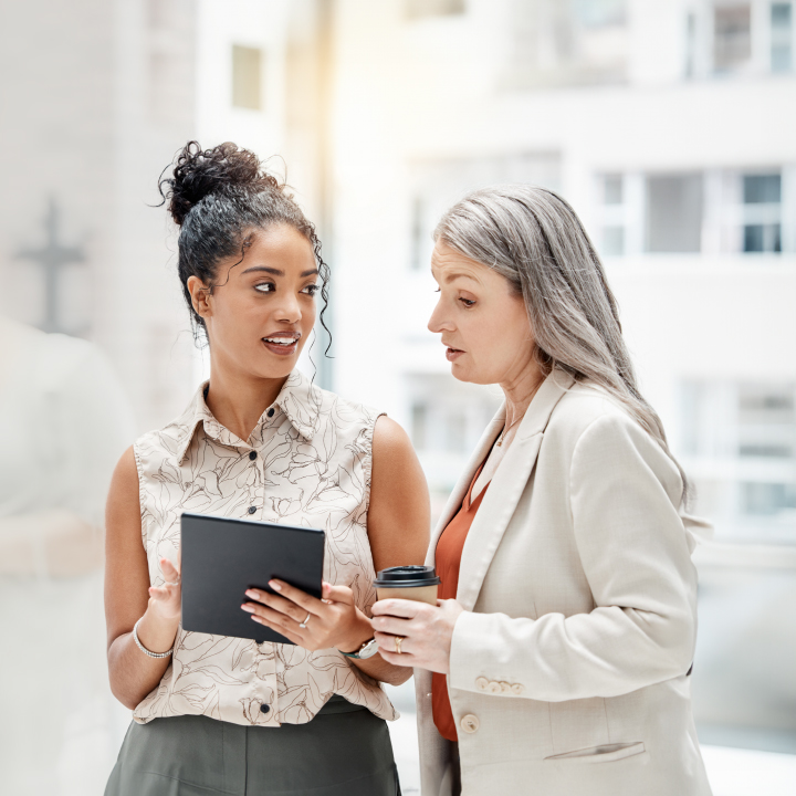 Dos ejecutivas charlan en un ambiente corporativo externo; una sostiene una tablet y la otra una taza de café, mientras discuten algo atentamente.