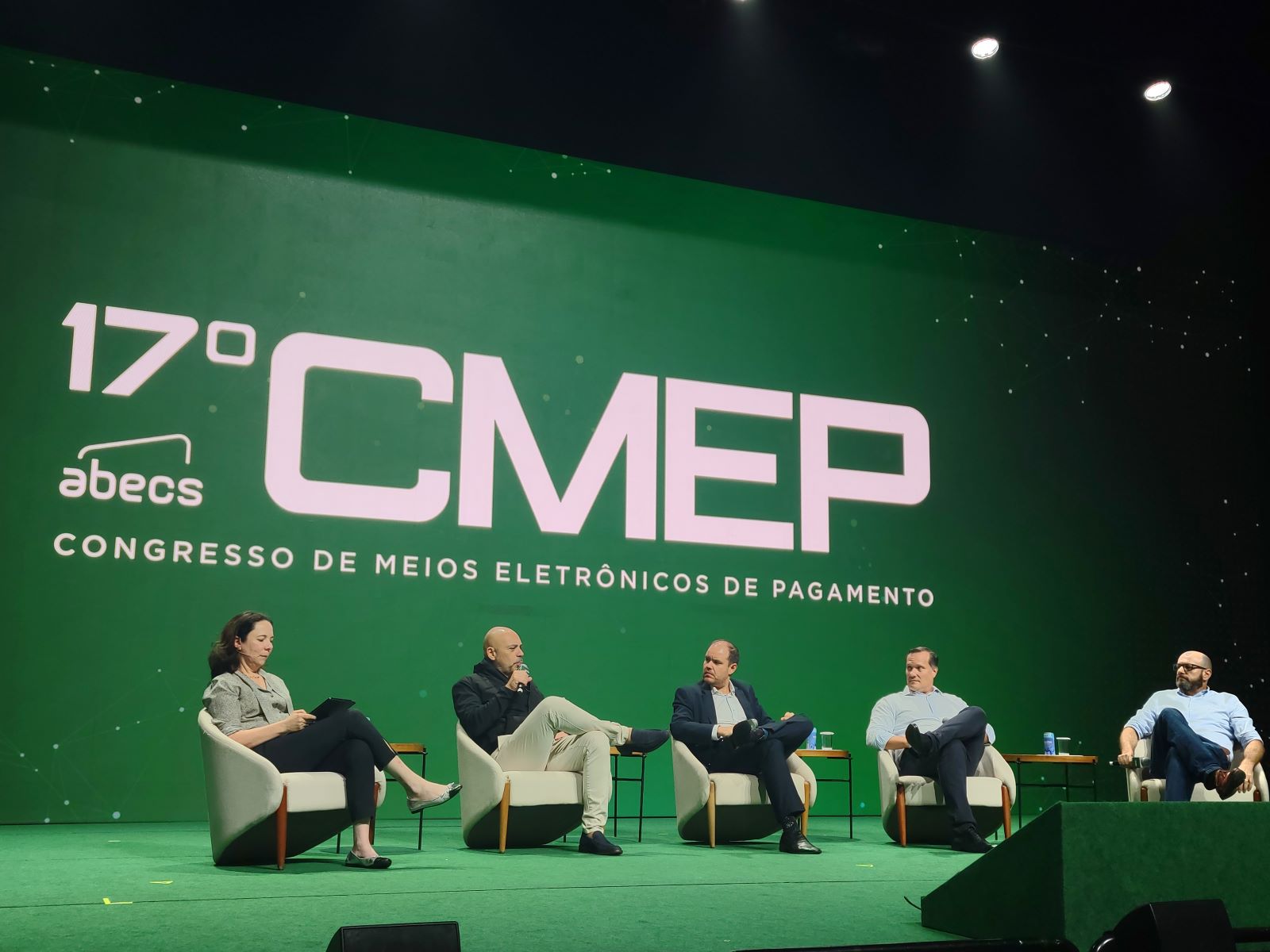 Quatro pessoas estão no palco que tem um telão com o logo do CMEP, evento sobre meios eletrônicos de pagamento, ao fundo.