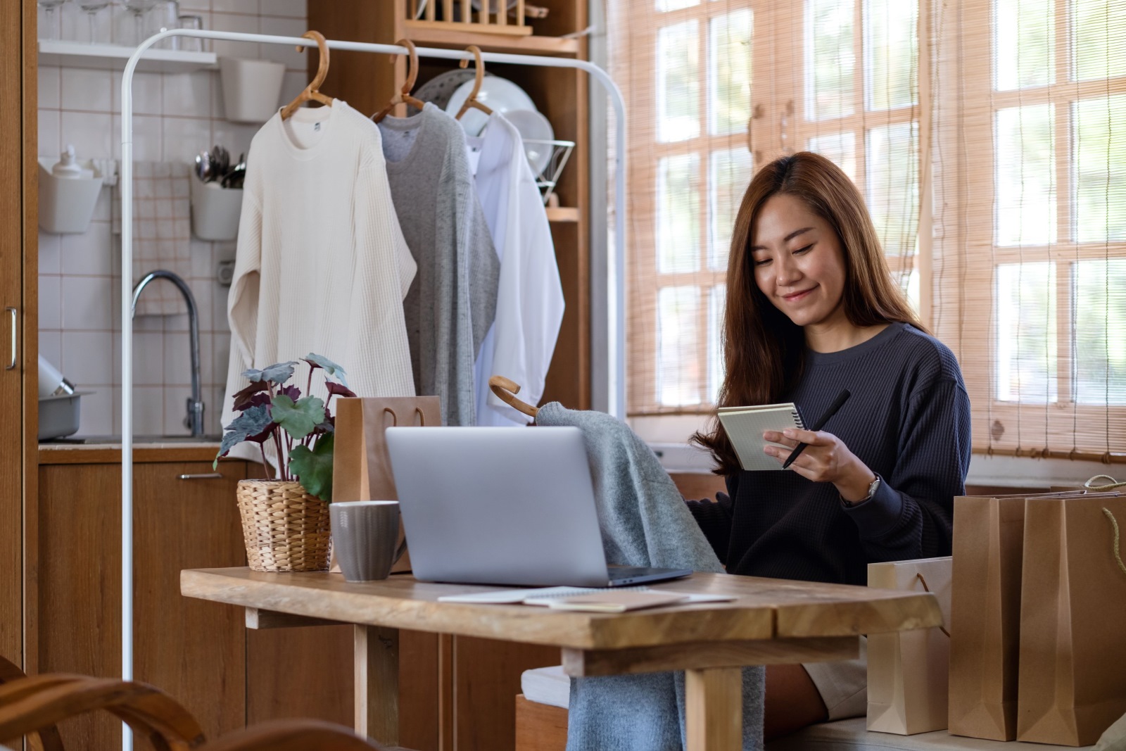 Mulher sentada em frente ao computador ao lado de uma arara de roupas administrando os negócios da sua marca online